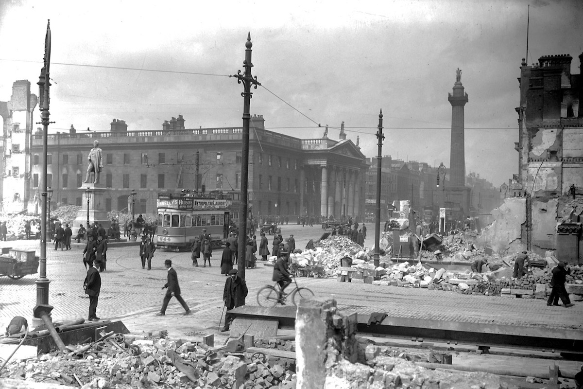 Schwarz-Weiß-Foto von Abbey Street und Sackville Street mit Statue von William Smith O'Brien, dem General Post Office und Nelson's Pillar. Man sieht Trümmerberge, Fußgänger, ein paar Autos und eine Trambahn.