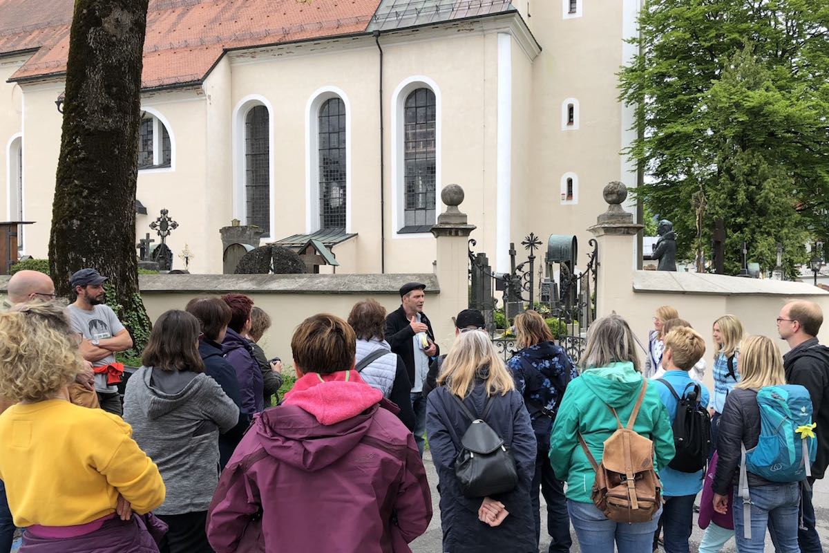Oliver Pötzsch stands in front of the cemetery wall and explains the origin of the Passion play to the group