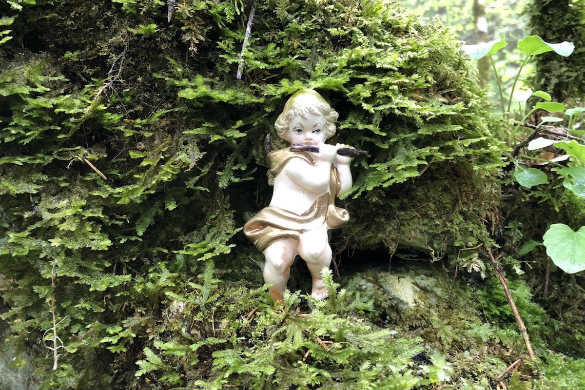 White figure of an angel with a flute standing in an area overgrown with moss