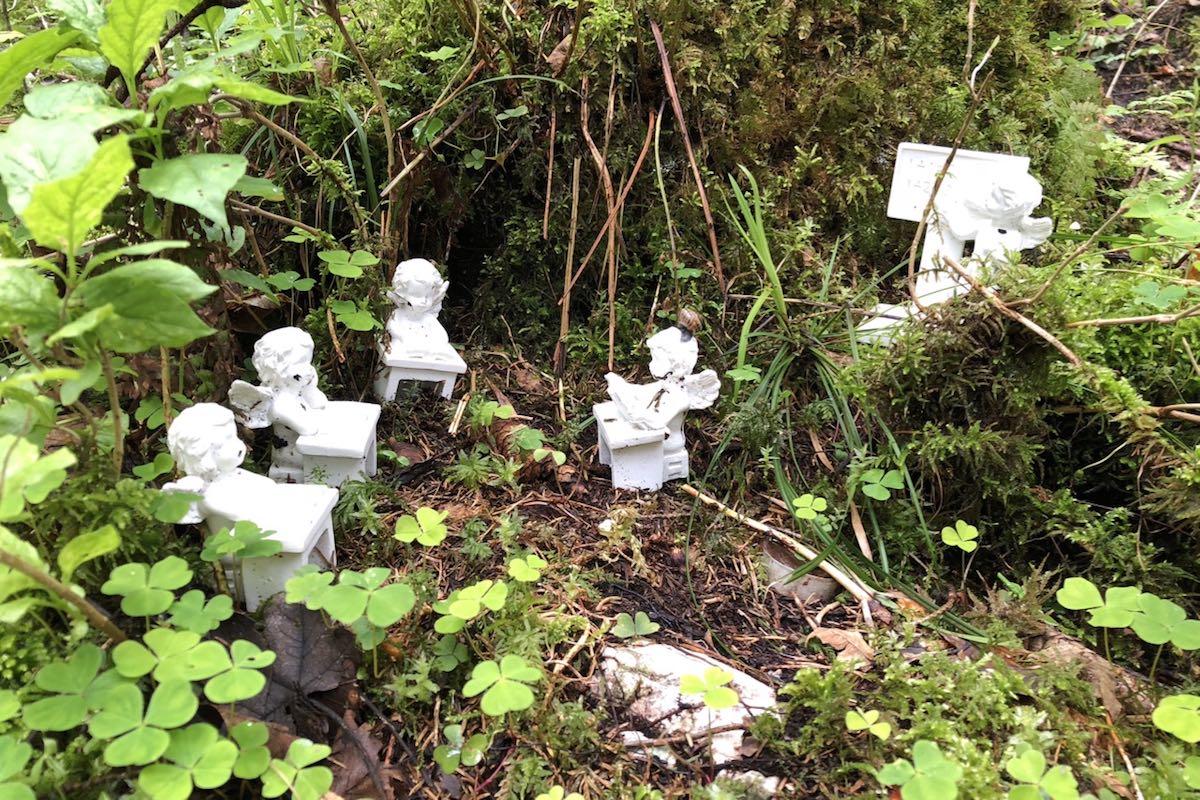 Four white figures of angels sitting at their little tables and one standing at a board in an area with shamrocks and moss